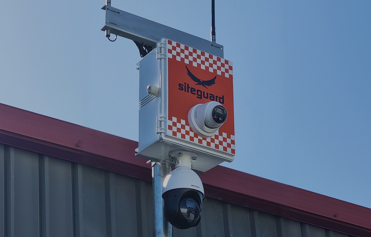 Siteguard security camera setup with two surveillance cameras mounted on a metal pole against a building and clear blue sky.